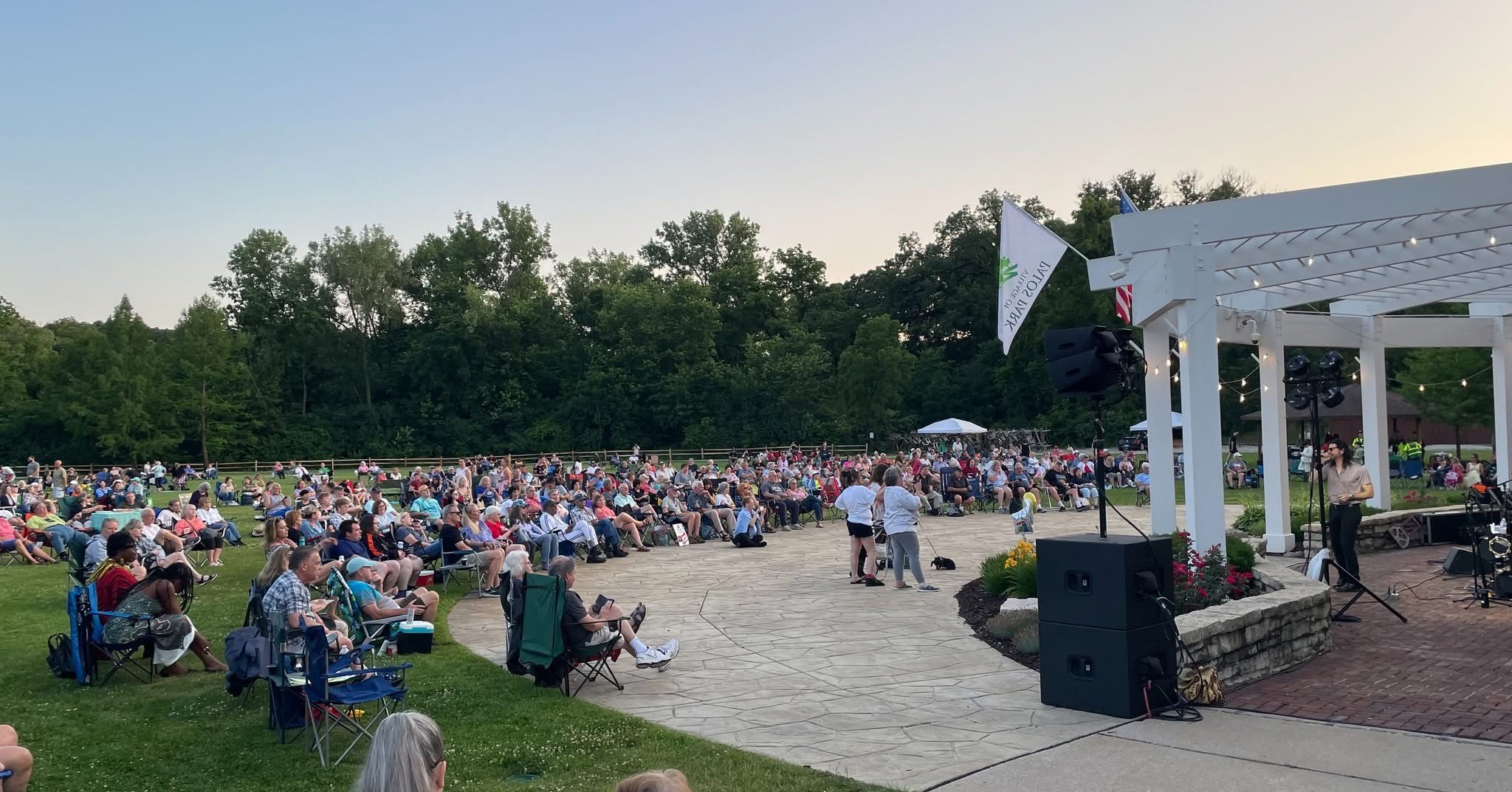 people in lawn chairs watching a performance in a white column bandshell